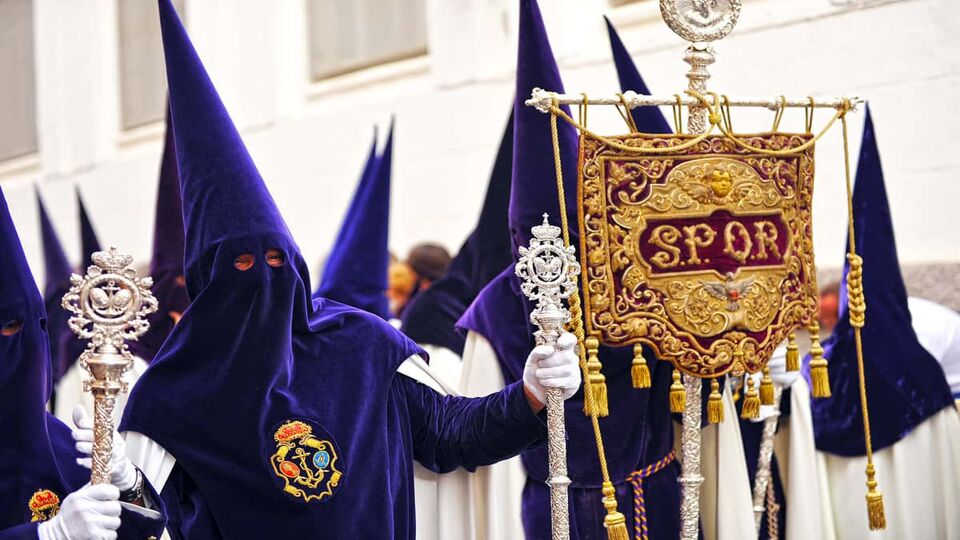 People wearing purple pointed hoods and white robes carry a flag tapestry
