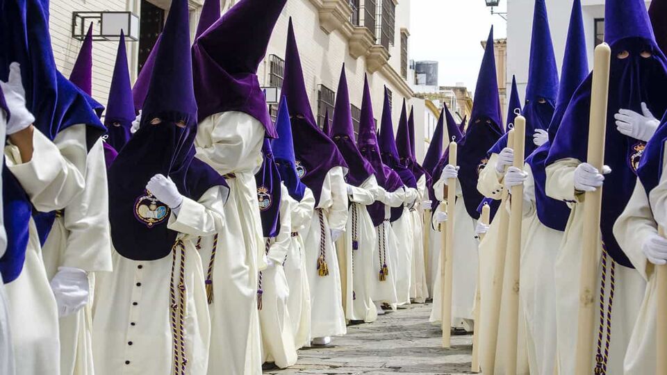 Long line of people dressed in white robes with pointed purple hoods that cover their faces