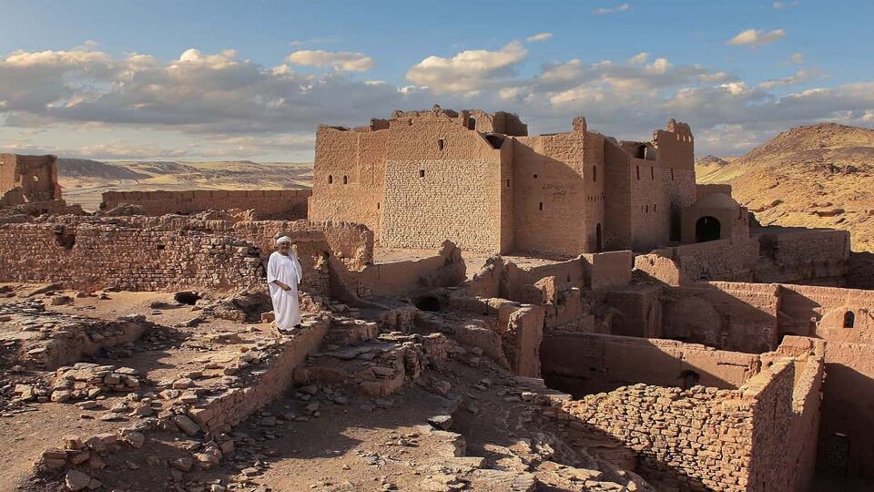 The old and dilapidated St. Simeon's Monastery in Upper Egypt. An Egyptian stands in front of the ruin in a white robe and a turban. It's late afternoon. A blue sky with clouds.