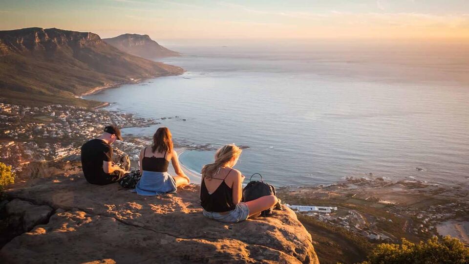 Young man and women sitting on the rock on top of Lion's head mountain look at the Atlantic Ocean and Cape town city view in South Africa