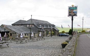 Jamaica Inn. Pub forecourt and sign. Outside tables. Cornish, American and UK flags. Moor in background. location for book by Daphne Du Maurier