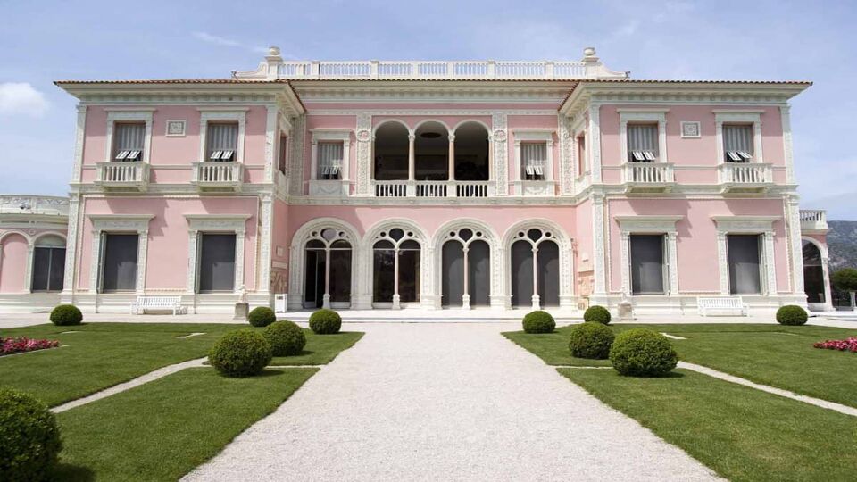 Walkway flanked by lawn leading to a large and ornate pink villa