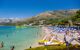 Sunny landscape side view of many people sunbathing on a beach with buildings and mountains in the background in Cavtat