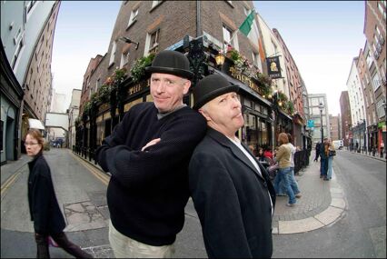 Two men standing back to back wearing black jackets and black fedoras on the streetpulling