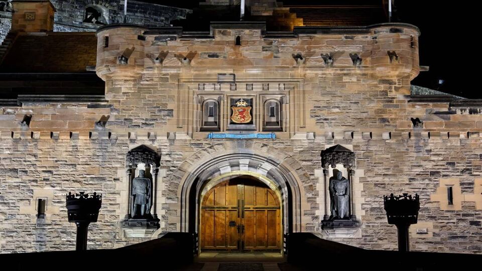 A photo showing the entrance of Edinburgh Castle at night where the castle is illuminated by lights. The wooden door is arched with metal accents around and there are a statue embedded into the wall on both sides of the door. The castle appears symmetrical with the silhouette of a torch on the left and right in the foreground.