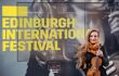 A photo of a Nicola Benedetti standing with her violin in front a large advertisement of herself for Edinburgh International Festival