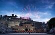 A far view of a firework show appearing behind a building. The Royal Edinburgh Military Tattoo is seen performing on the floor ground.