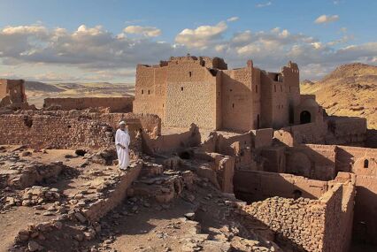 The old and dilapidated St. Simeon's Monastery in Upper Egypt. An Egyptian stands in front of the ruin in a white robe and a turban. It's late afternoon. A blue sky with clouds.