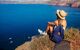 Woman wearing a sunhat sits on the edge of a rock and looks out over the caldera