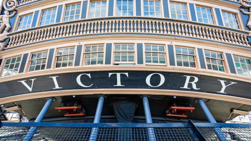 Close up of stern of HMS Victory, Lord nelson's flagship, on display at Portsmouth Dockyard. The striking exterior of small men in statues, attached to both sides of the ship, with 2 rows of windows in a slight curved structure. The word 'Victory' can be seen at the bottom of the ship, pained in white against the black ship.