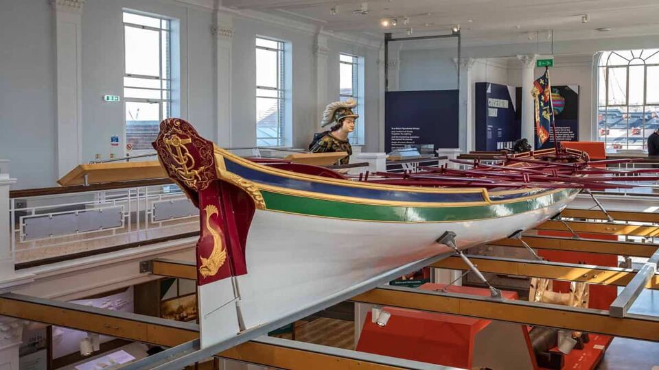 The royal barge displayed in a room. The boat is painted white with green, red and yellow accent detailing around. This barge took Lord Nelson's coffin from Greenwich to St Paul's Cathedral on display in Portsmouth Dockyard, Hampshire, UK on 29 September 2021