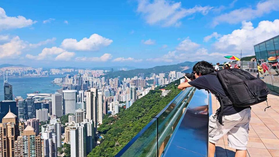 Tourist takes photo over the edge of the Peak Tower, with the harbour and skyline visible in the background