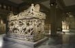 Intricately-carved sarcophagus in a darkly-lit museum gallery with other ancient artefacts in the background
