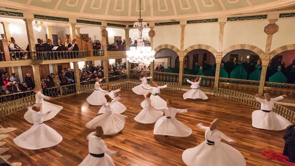 A large group of dancers spin on a wooden dance floor in a grand hall as musicians play on an upper balcony and guests spectate below