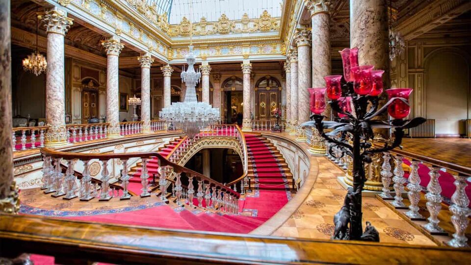 Red-carpeted, ornate staircase in palace with chandelier