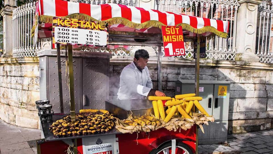 A street vendor selling whole grilled ears of corn and roasted chestnuts from his cart trimmed with a red and white awning