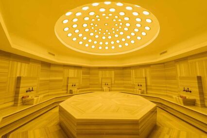 Interior of a marble, Turkish bathhouse which is hexagonal and cast under yellow light from the skylights above