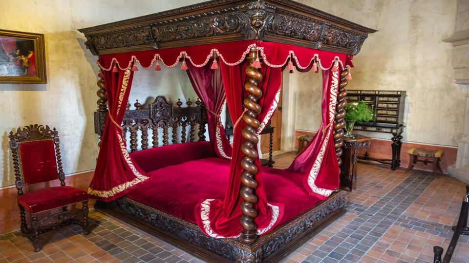 A bedroom inside the chateau in the style of the middle ages with a red canopy bed and a dark brown polished desk in the centre of the room. Along with a large medieval fireplace.