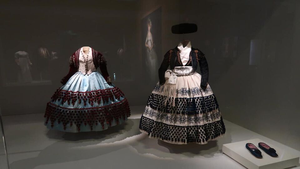 Two elaborate women's costumes on display behind glass at the Costume & Fashion Museum. Both include petticoated skirts and buttoned blouses with long sleeved cropped jackets. One has a hat suspended above it, with a pair of black slip on shoes placed next to it.