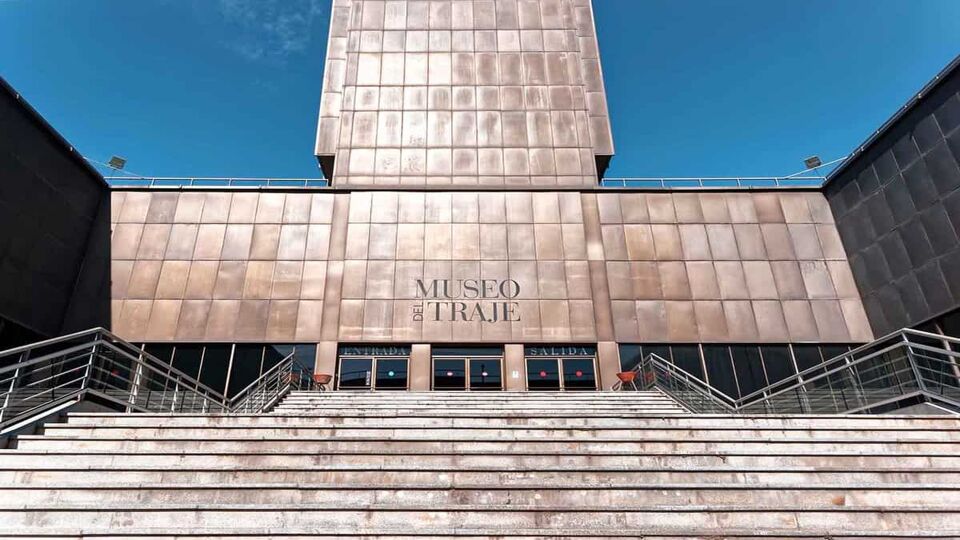 Main entrance to the Costume Museum or Museo del Traje in Madrid, Spain. The building is imposing and almost looks to be made of hammered plates of grey-red metal. There are steps leading up to the entrance, and the sky is clear and blue.