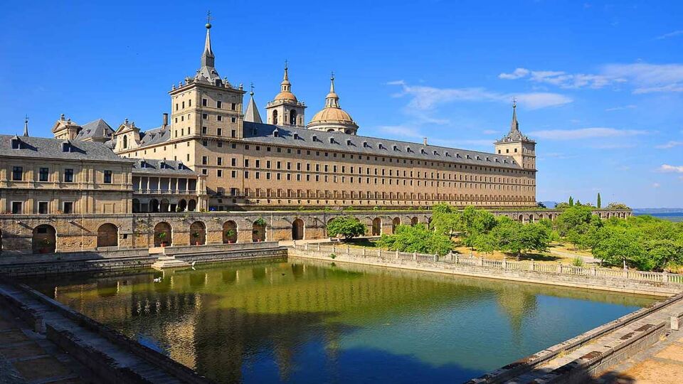 El Escorial Palace. The yellowish stone building is Herrerian style architecture, made up of a rectangular base with many small and narrow windows, with domes and spires rising from the centre. The roof is grey slate. In the image a large rectangular pond is in the foreground, and the sky is blue with wispy clouds.