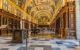 Inside the royal library of El Escorial Palace. The floors are chequered white, grey, and black marble. The lower half of the wall is panelled wood with hanging oil portraits, and the upper half and ceiling has bright yellow gold moulding that splits the area into panels. Each panel has painted artwork within it, from the upper half of the walls curving up onto the arched ceiling.