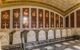 An image of the burial vault in El Escorial Palace. Stone sarcophagi line up in a row, raised from the ground by a dark grey marble plinth. Each sarcophagus is pale stone, with latticework etched along the top, and a banner with writing above it carved into the foot of the stone coffins (which is facing towards the viewer). Above each sarcophagus is a stone cross, and the wall above each coffin is dark red stone engraved with their name and decorated by their crest in full colour.