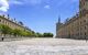 A view from a courtyard within El Escorial Palace. Several tourists are dotted around the large courtyard taking pictures, and appear small and far away. A palace building sits on each side of the frame, yellowish stone with many small windows. There is a line of small trees running through the centre of the courtyard.