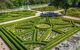 A garden in El Escorial Palace viewed from a high angle. In the centre is a circular fountain. A pale terracotta coloured path extends from the fountain in four directions, forming a rectangle in each corner. The corner rectangles have ornamental hedges that form the pattern of a circle, with triangles extending from it to fill the rectangle.