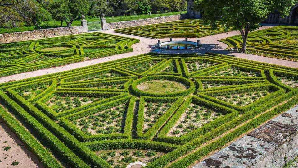 A garden in El Escorial Palace viewed from a high angle. In the centre is a circular fountain. A pale terracotta coloured path extends from the fountain in four directions, forming a rectangle in each corner. The corner rectangles have ornamental hedges that form the pattern of a circle, with triangles extending from it to fill the rectangle.