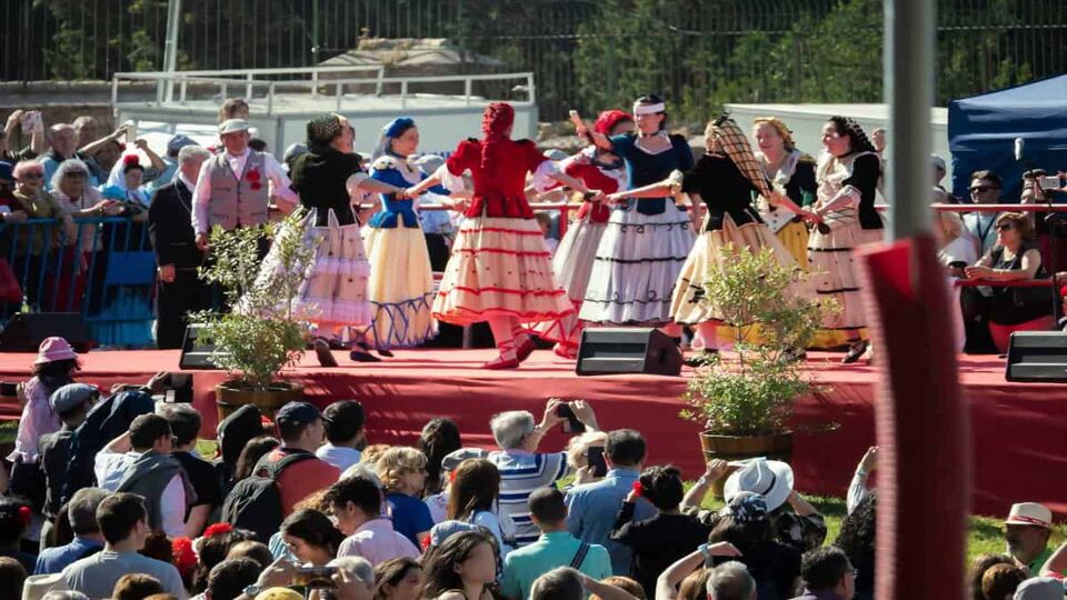 A folk dance group performs on a raised platform covered in red cloth, for a crowd of festivalgoers. The performers are wearing colourful skirts and dancing in a circle while holding hands.