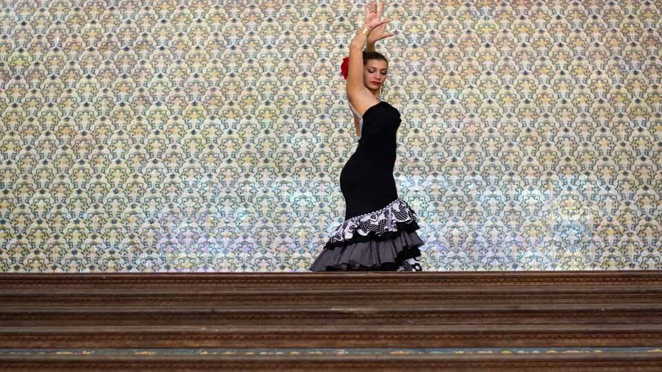 A flamenco dancer wearing a black flamenco dress poses with her arms above her head at the top of a series of steps. She is wearing a red flower in her hair.