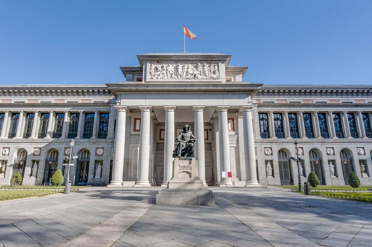 The grand exterior of the Prado Museum. Made of white stone with multiple pillars, and a carved stone mural set at the top of the entrance building, there is a flag flying atop it. A large statue of a figure cast in dark metal sits in front of the entrance.