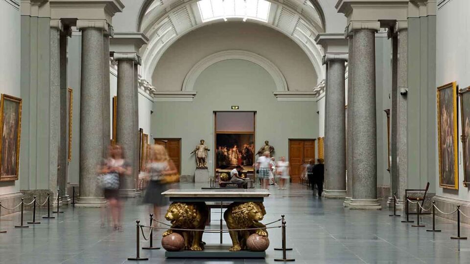The main gallery of the Prado Museum, on the ground floor. There are curved ceilings, skylights, and stone pillars and floors. Visitors walk around, and there are oil paintings hung on the walls. A low sculpture is in the centre of the room, sectioned off by rope.