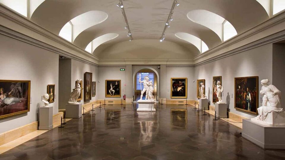 The main gallery of the Prado Museum, on the ground floor. There are lit alcoves in the ceiling, and a shiny wooden floor. The room contains white marble statues and framed oil paintings