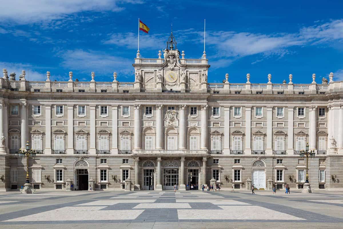 The exterior of the Royal Palace of Madrid. It is a grand building made of pale grey stone, with a Spanish flag flying atop it. The courtyard in front is made of dark and light grey stone, and the sky is blue with wispy clouds.