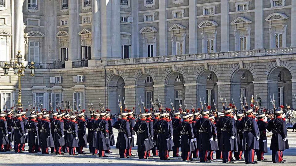 Guards line up outside the Royal Palace of Madrid, ready to enact the changing of the guard ceremony. They wear blue uniforms and hats, white gloves, and carry flags.