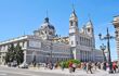 Almudena Cathedral. A cathedral, with a central dome, pillars, and decorative elements. Pedestrians walk in the square in front of it.