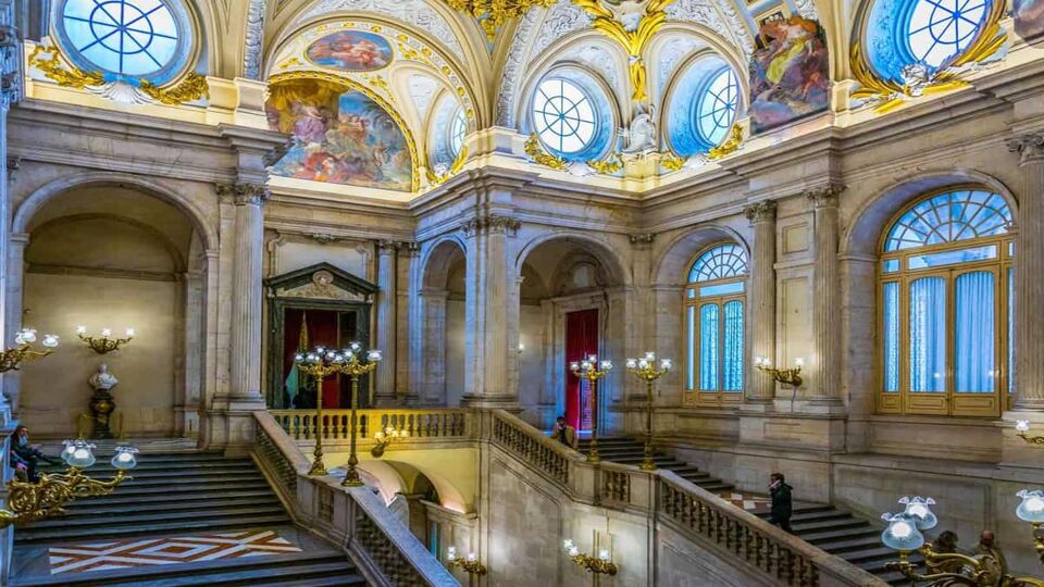 View of the main decorated lobby with a staircase in the Royal Palace of Madrid. There are circular windows, patterned tile floors, and sconces with glass lamps.