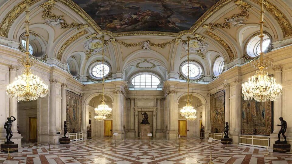The Neoclassical Columns Room (Salon de las Columnas) in the Royal Palace of Madrid. The floor is white and red marble, with tiles places in a pattern. There are circular windows, gold moulding over white painted or white stone elements, a fresco painted ceiling, and chandeliers.