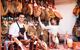 A smiling butcher stands in front of two racks of hanging Jamon Iberico. A colleague reaches up behind him to take one down for a customer,