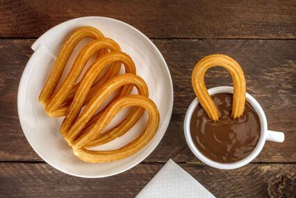 An overhead photo of churros on a white plate. One churro has been half dipped into a mug of hot chocolate.