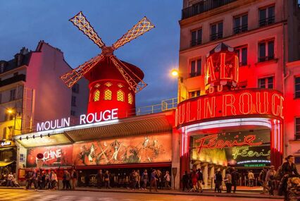 Front view of the Moulin Rouge on a evening with bright neon red lights and a long queue