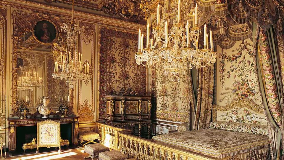 Inside view of a royal chamber where there are floral patterns on the wall with gold colour linings, a huge chandelier and a bed.