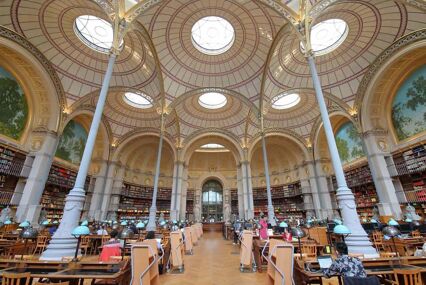 Unidentified people visiting the National Library of France. There are tall skinny podiums reaching the high ceilings, where there are round circular windows in the ceiling.