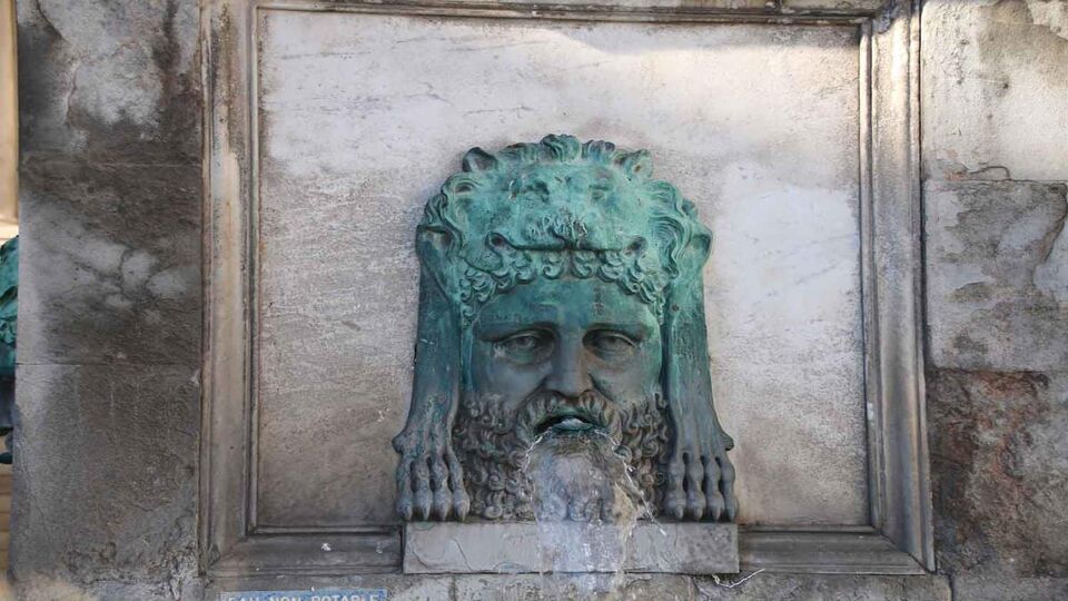 A close up image of a green water spout in the shape of a man's head at the base of the Arles Obelisk