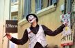 An actor with a painted face, waistcoat, and a bowler hat, walks on stilts and holds fliers to advertise his performance