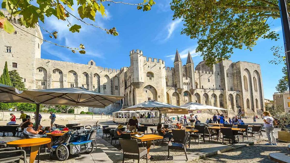 Tourists enjoy a meal at a sidewalk cafe in front of the medieval Pope's Palace in the Provence region of Avignon France as a tourist tram drives by on a sunny day