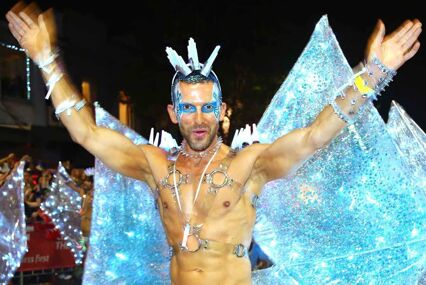 A topless man, wearing vibrant blue wings, holding his hands up.