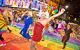 A group of people in vibrant costumes with the LGBTQ+ flag painted on the street. In the foreground there is a drag Queen in a short red dress striking a pose.